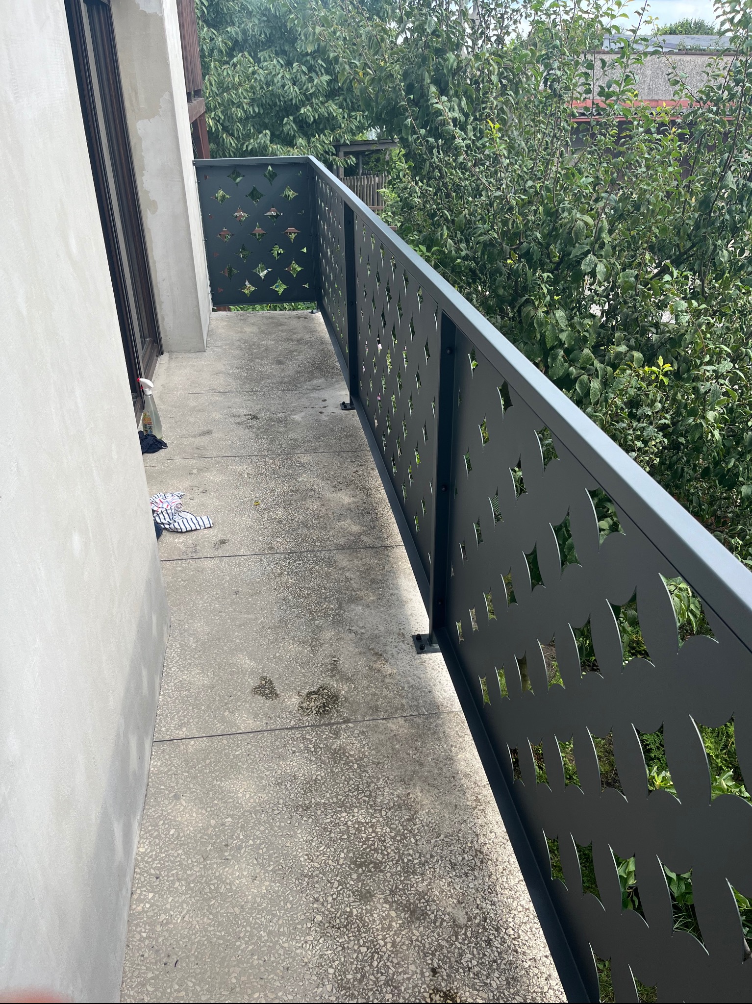 Nowoczesna, szara balustrada balkonowa z geometrycznym, ażurowym wzorem na tle zieleni ogrodu, widok z boku, na podłodze leżą szmaty i butelka z detergentem.