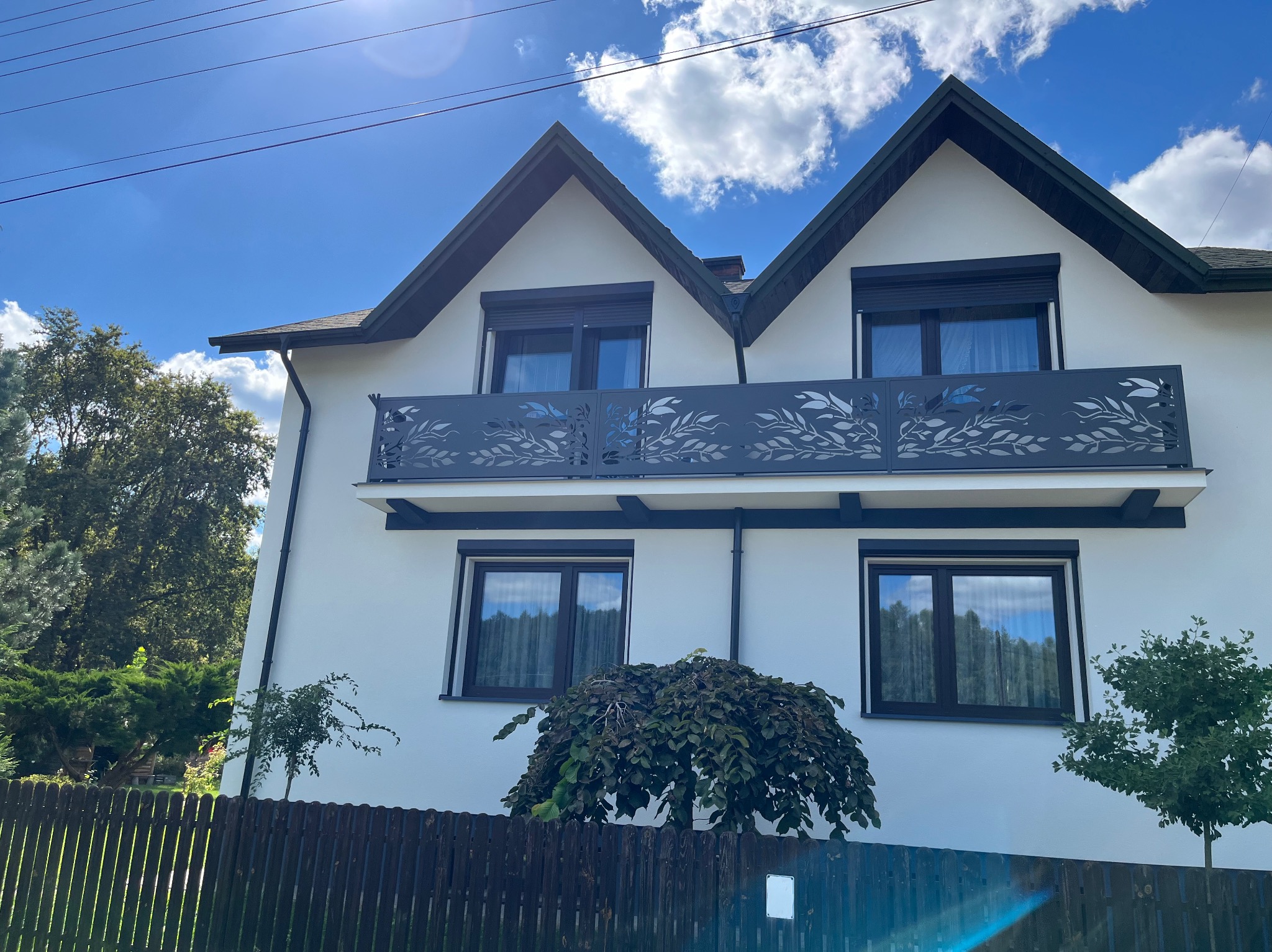 Biały dom z balkonem z metalową balustradą o wzorze liści, widok z poziomu ogrodzenia.