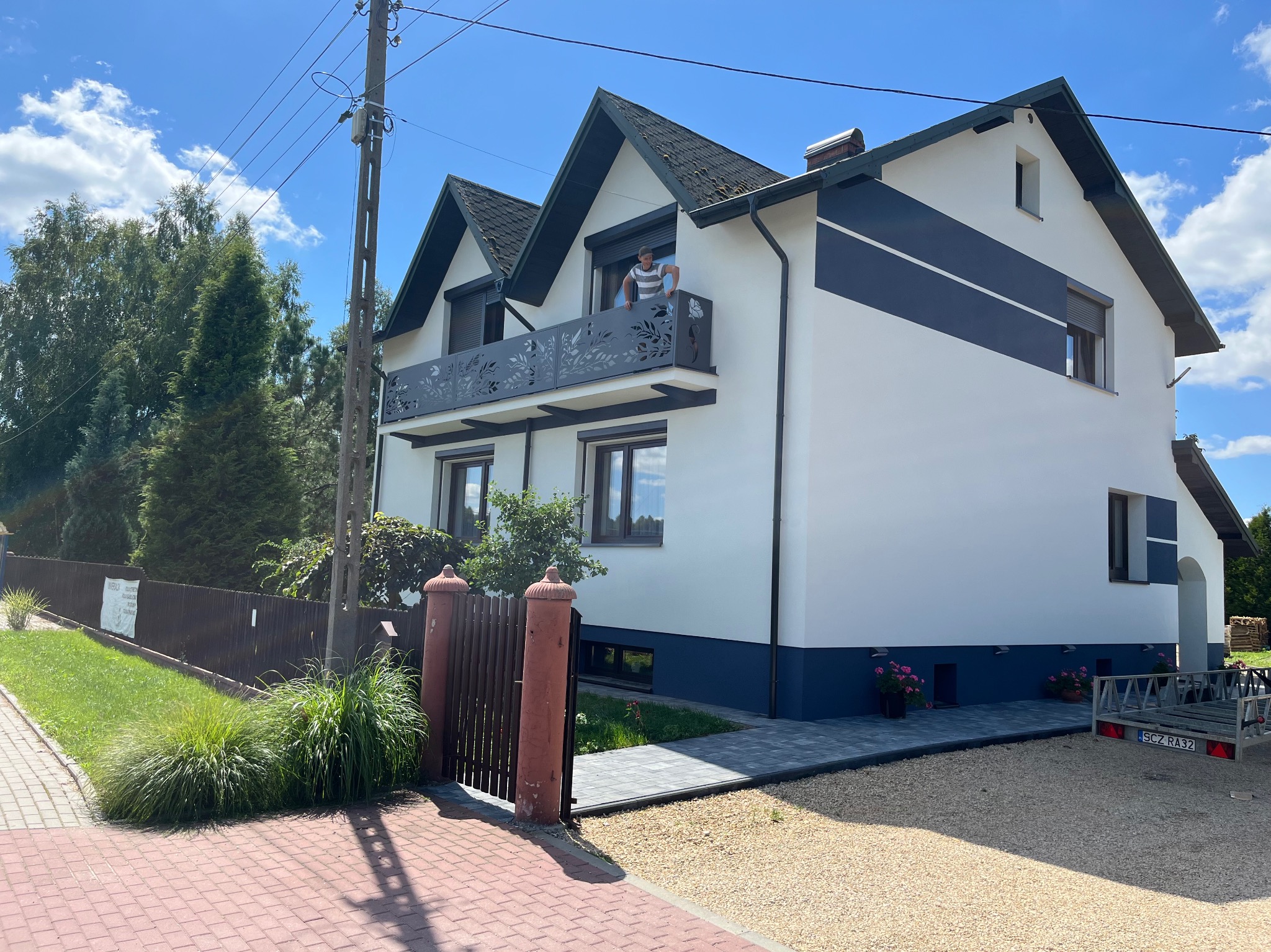 Nowoczesny dom z białymi ścianami i dekoracyjnym, metalowym balkonem z motywem roślinnym, osoba opiera się o barierkę balkonu, widok z zewnątrz na podwórze z kostką brukową i ogrodzeniem.