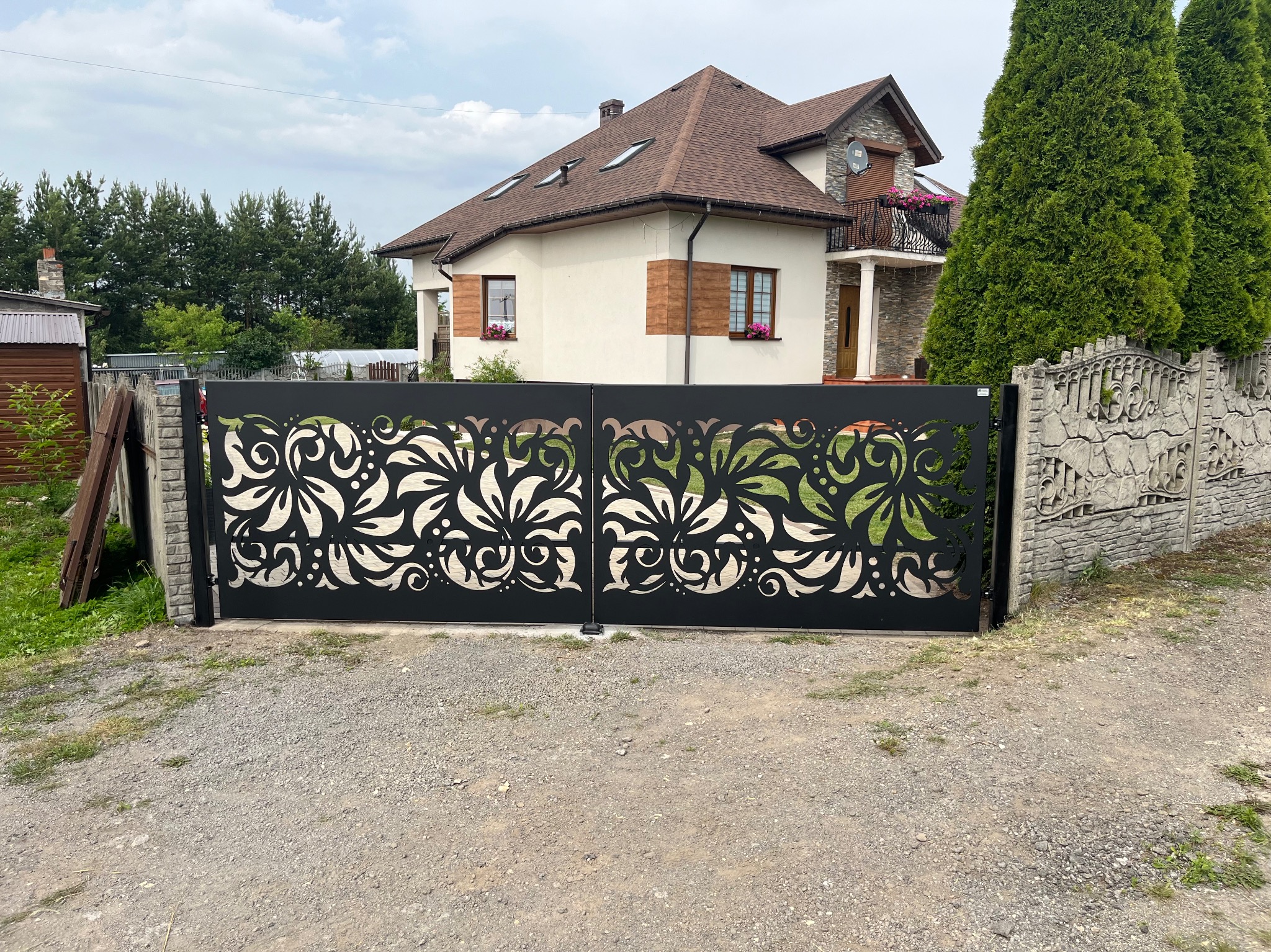 Czarna, dwuskrzydłowa brama wjazdowa z ażurowym, roślinnym wzorem na tle domu z beżową elewacją i brązowym dachem.