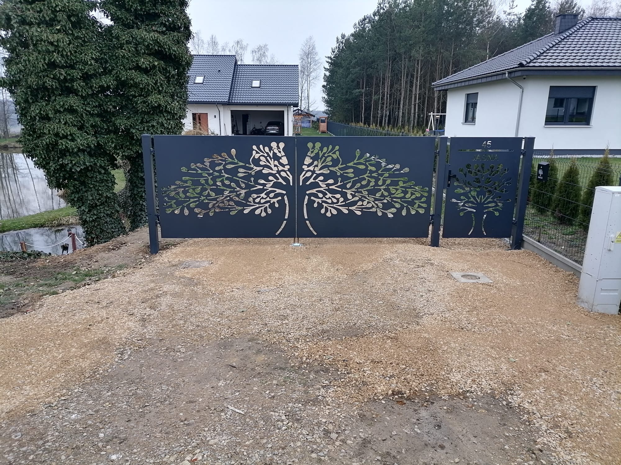 Nowoczesna, grafitowa brama wjazdowa z laserowo wycinanym motywem drzewa, na tle nowo wybudowanego domu i drzew.