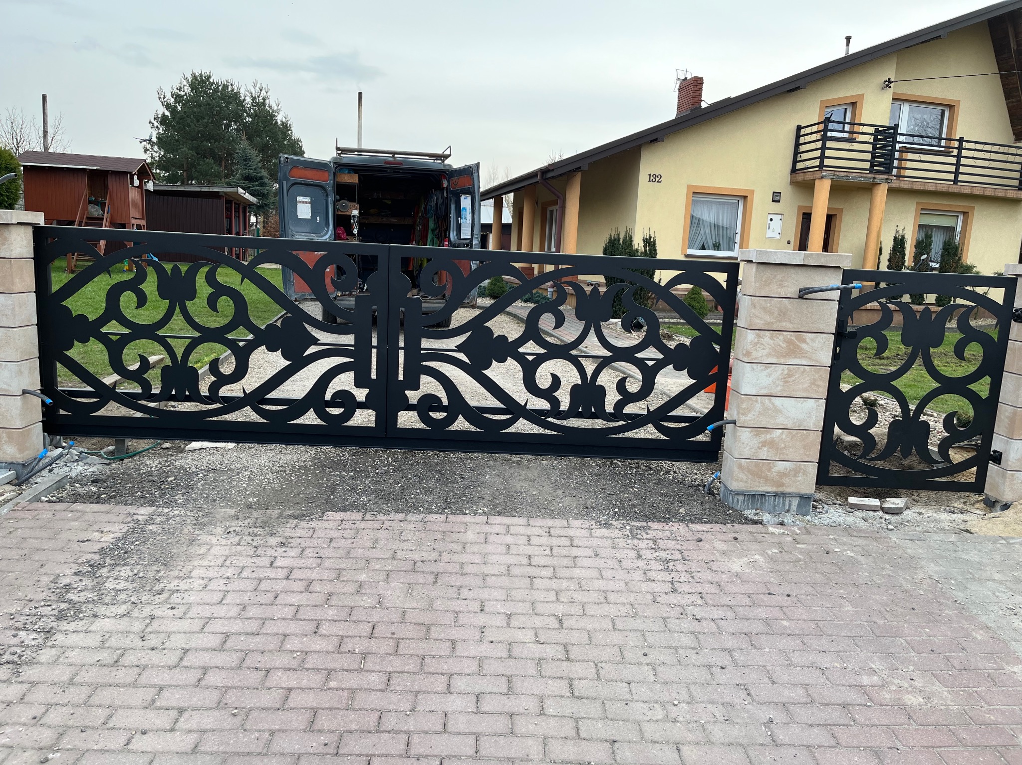 Czarna, ozdobna brama wjazdowa z kutymi elementami, częściowo zamontowana na posesji przed domem z numerem 132, z samochodem dostawczym w tle.