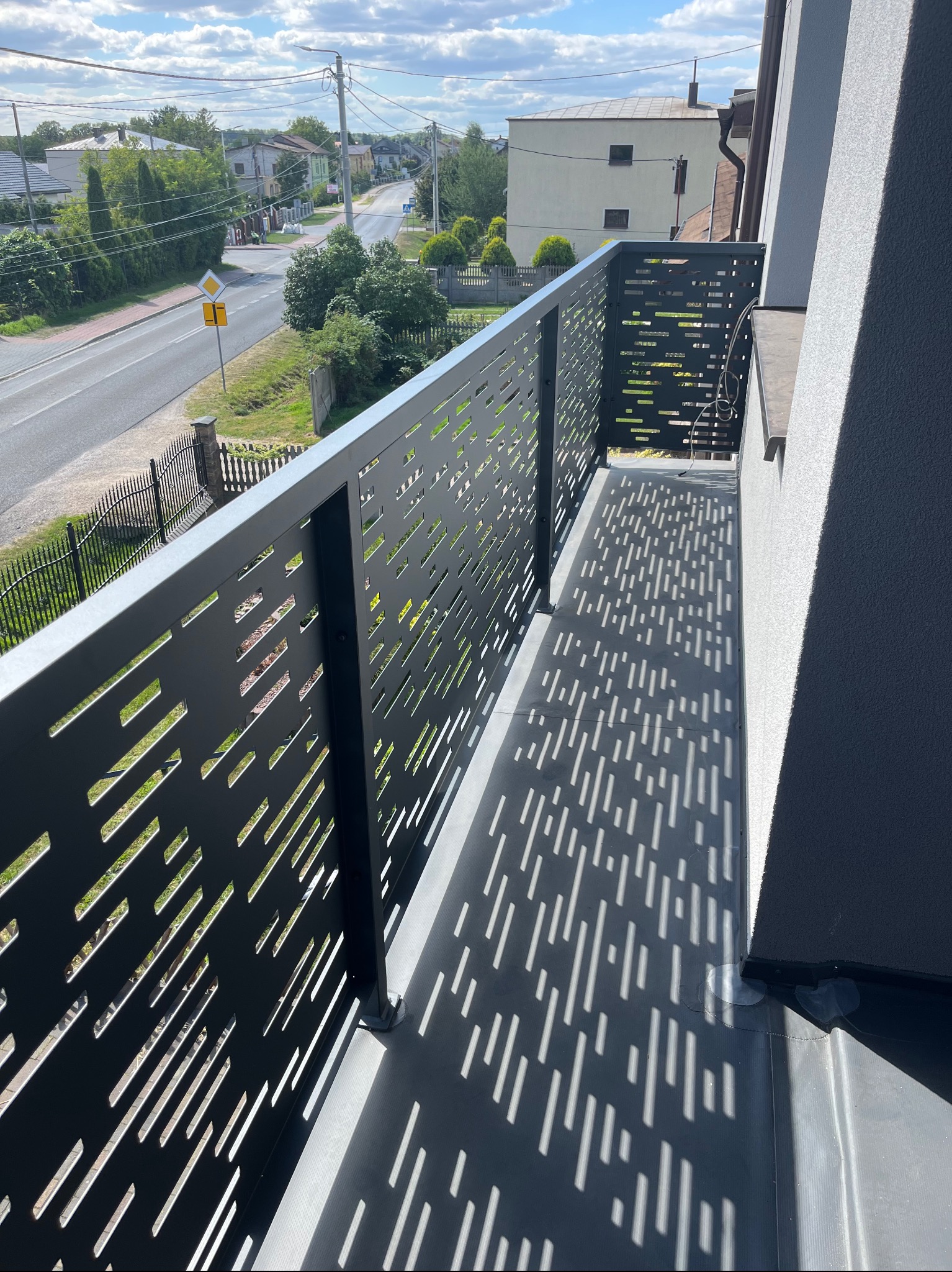 Nowoczesna, czarna balustrada balkonowa z laserowo wycinanym wzorem, rzucająca cienie na podłogę balkonu. W tle widok na ulicę i zabudowania.