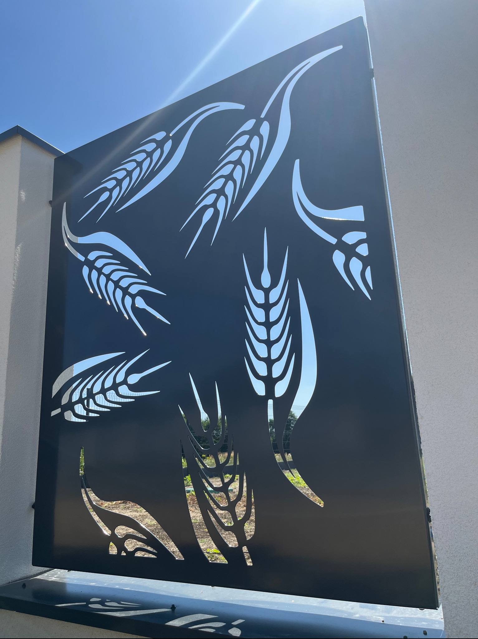 Czarna brama wjazdowa z wyciętym wzorem kłosów zboża, widziana pod kątem, z jasnym niebem w tle. Nowoczesny design z motywem roślinnym.