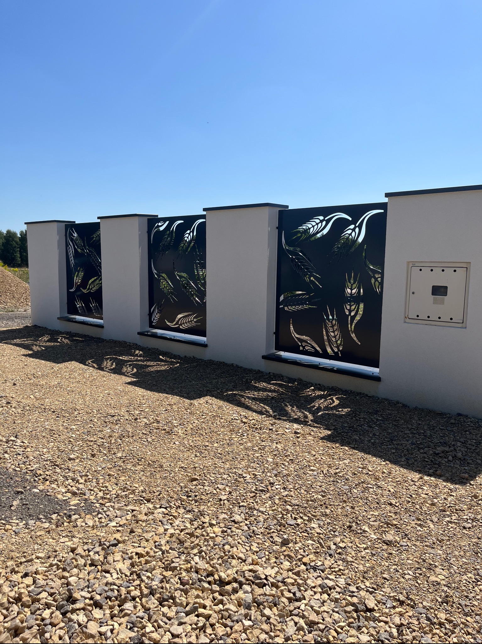 Nowoczesne ogrodzenie panelowe z motywem roślinnym, czarne panele między białymi słupkami, żwirowe podłoże, błękitne niebo w tle.