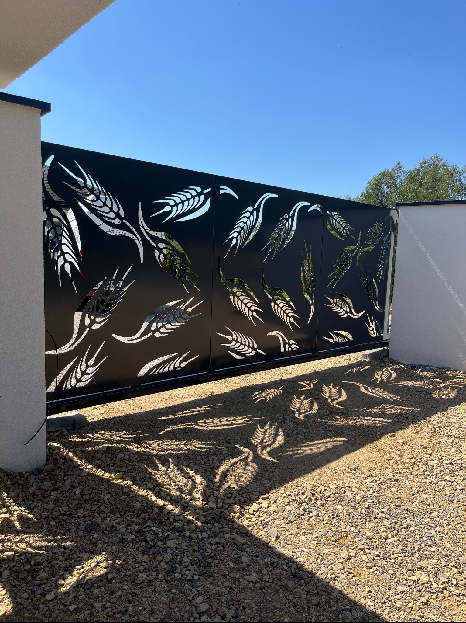 Nowoczesna, przesuwna brama wjazdowa z motywem roślinnym, rzucająca cienie na żwirowe podłoże w słoneczny dzień. Czarna brama z wycinanymi wzorami.