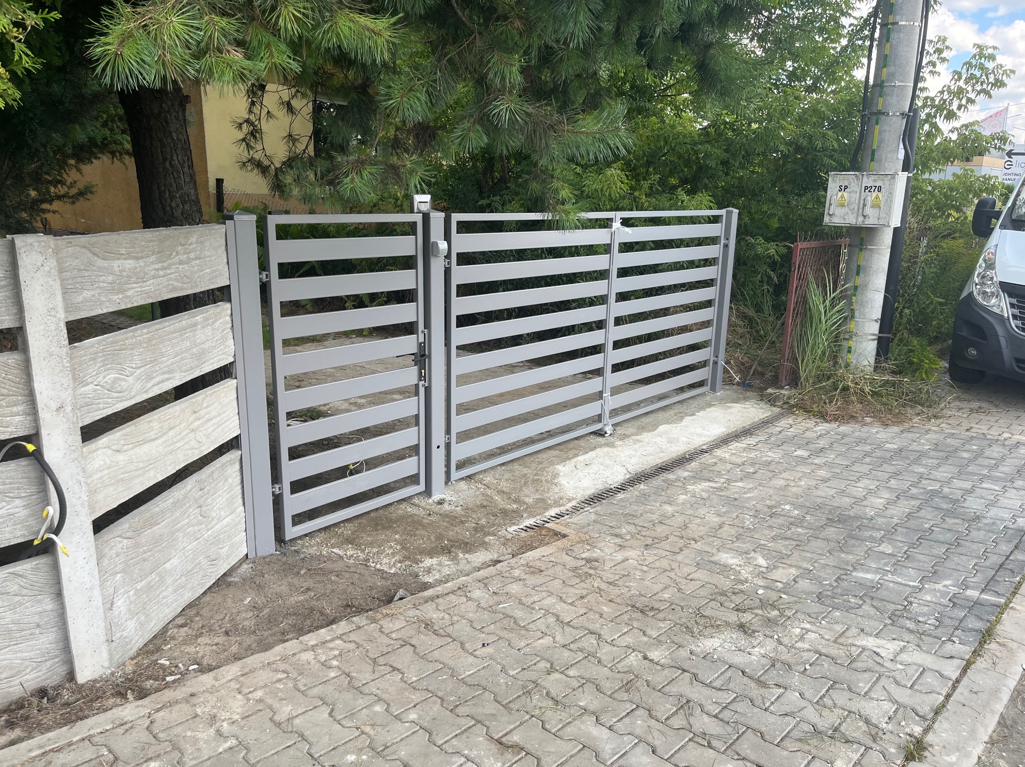 Otwarta, szara brama wjazdowa z poziomymi szczeblami, betonowy słupek imitujący drewno, kostka brukowa, zieleń w tle. Widok z zewnątrz posesji.