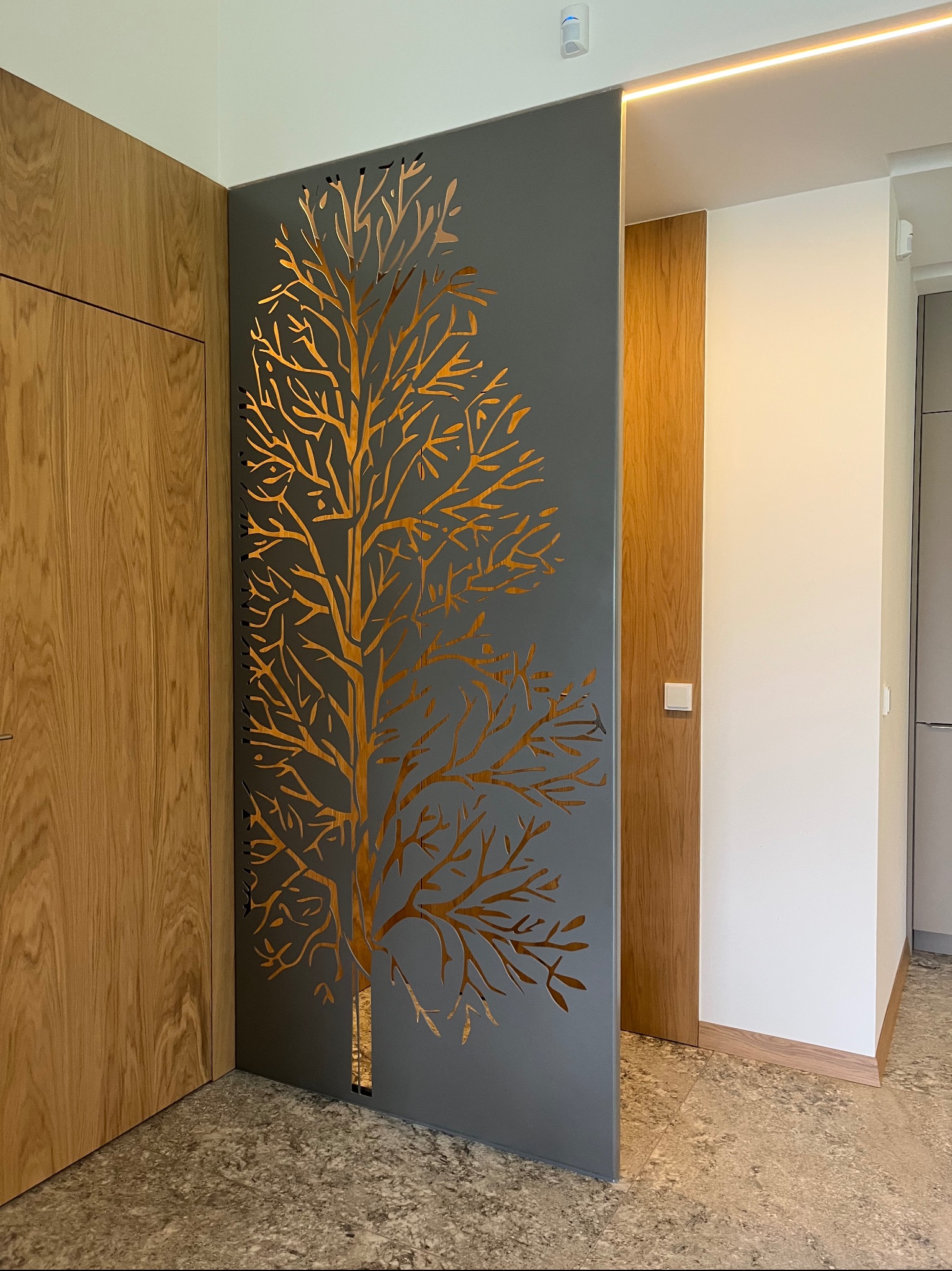 Metalowa ścianka działowa z wyciętym wzorem drzewa, oddzielająca przestrzeń w nowoczesnym wnętrzu z drewnianymi elementami i oświetleniem LED.