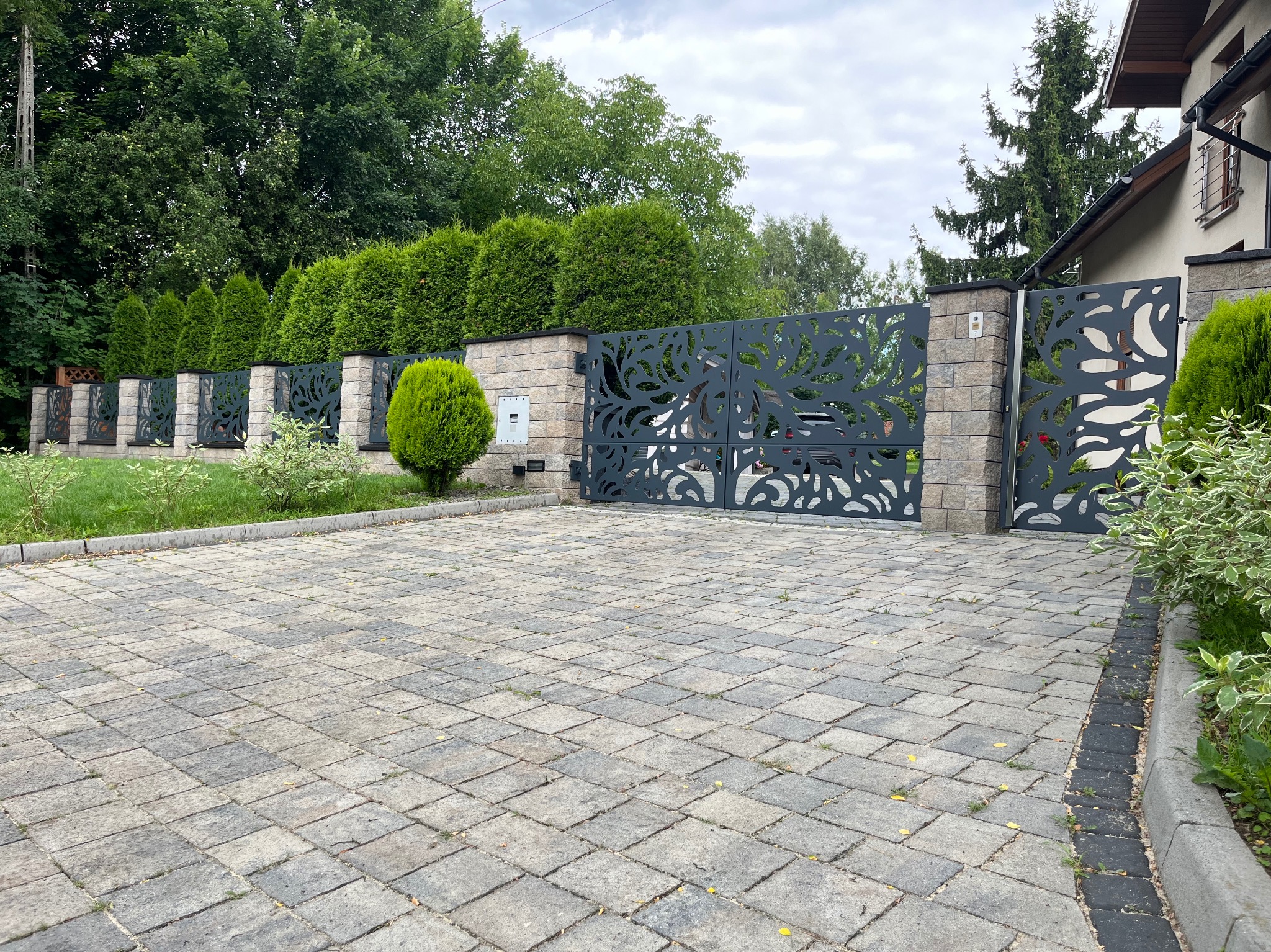 Nowoczesna, grafitowa brama wjazdowa z laserowo ciętym wzorem roślinnym, osadzona na murowanych słupkach. W tle żywopłot z tuj i kostka brukowa na podjeździe.