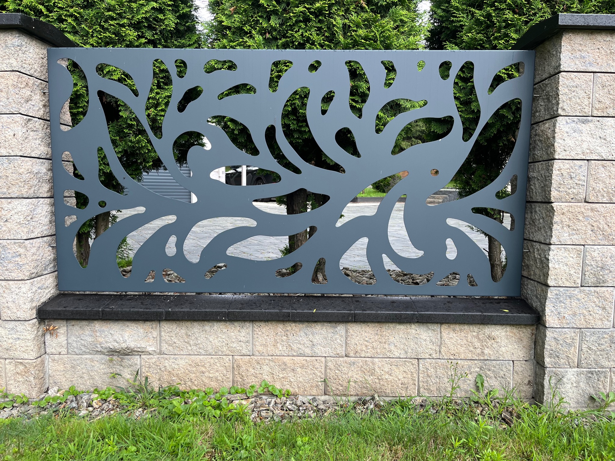 Nowoczesne ogrodzenie metalowe z dekoracyjnym wzorem wyciętym laserowo, osadzone na kamiennej podmurówce, z zielenią w tle.