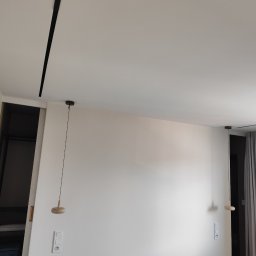 OLEH HRYNCHYSHYN - Minimalistyczne wnętrze: białe ściany, drewniane lampy wiszące i czarna listwa sufitowa. Widoczne włączniki i gniazdka. Elementy garderoby w tle.