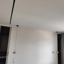 Minimalistyczne wnętrze: białe ściany, drewniane lampy wiszące i czarna listwa sufitowa. Widoczne włączniki i gniazdka. Elementy garderoby w tle.