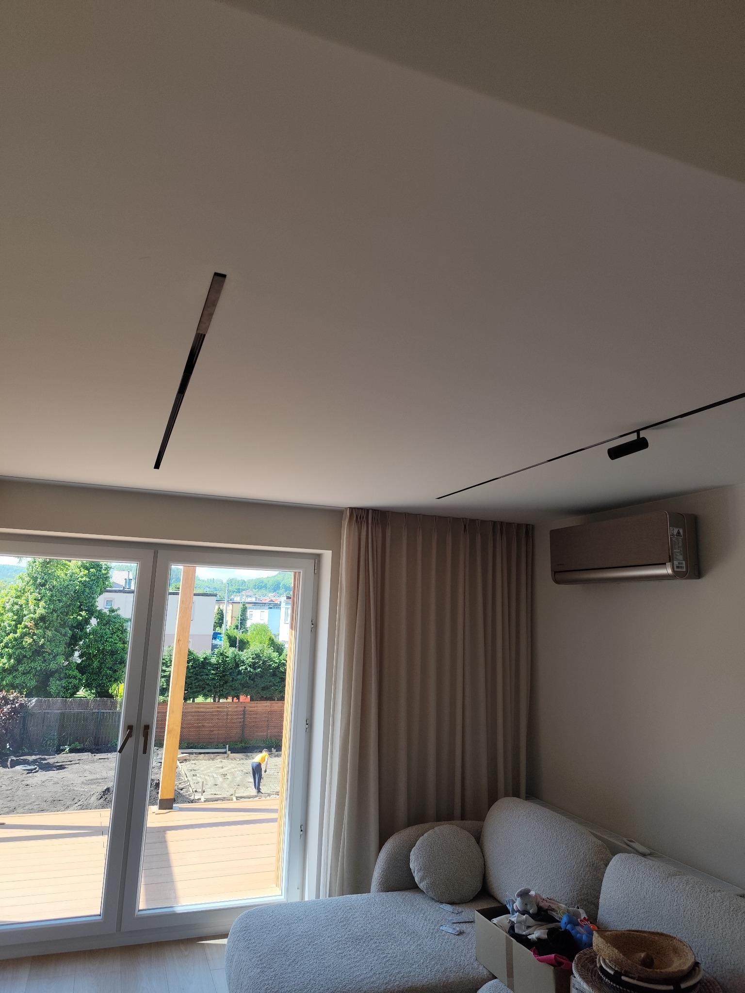 Nowoczesny salon z klimatyzacją, oświetleniem szynowym i dużym oknem z widokiem na ogród w trakcie budowy tarasu.
