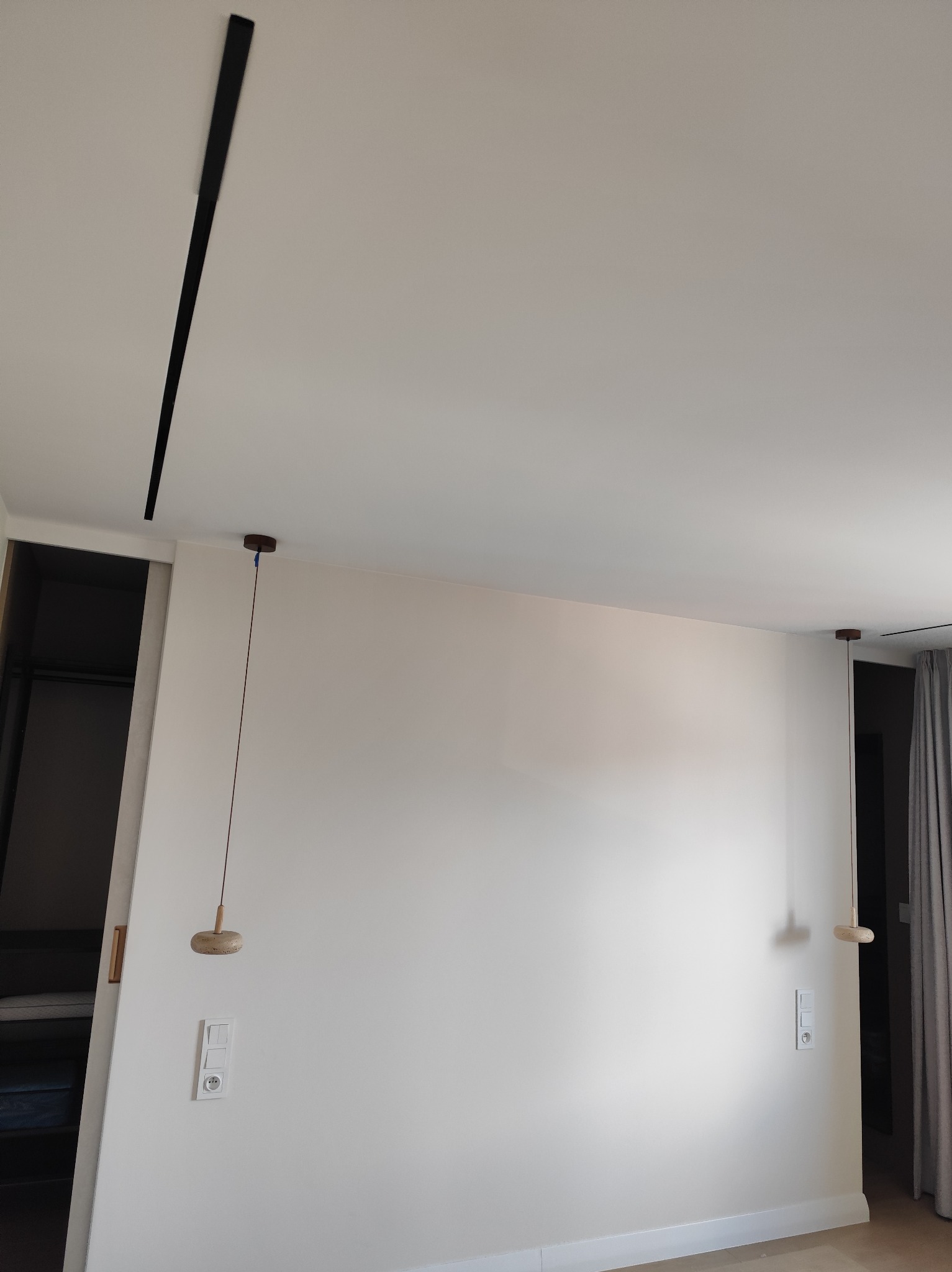 Minimalistyczne wnętrze z białymi ścianami, drewnianymi lampami wiszącymi i czarną listwą oświetleniową na suficie. Widoczne gniazdka elektryczne i fragment zasłony.