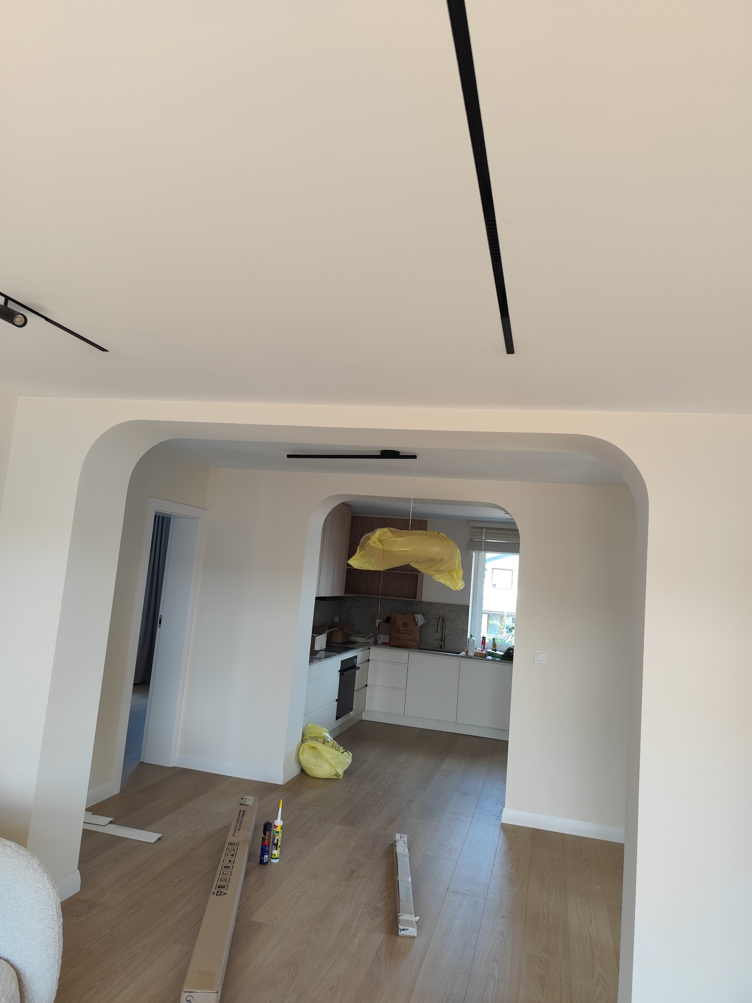 Jasne wnętrze po remoncie z drewnianą podłogą, widokiem na kuchnię przez łuk i czarnymi listwami oświetleniowymi na suficie. Na podłodze materiały budowlane.