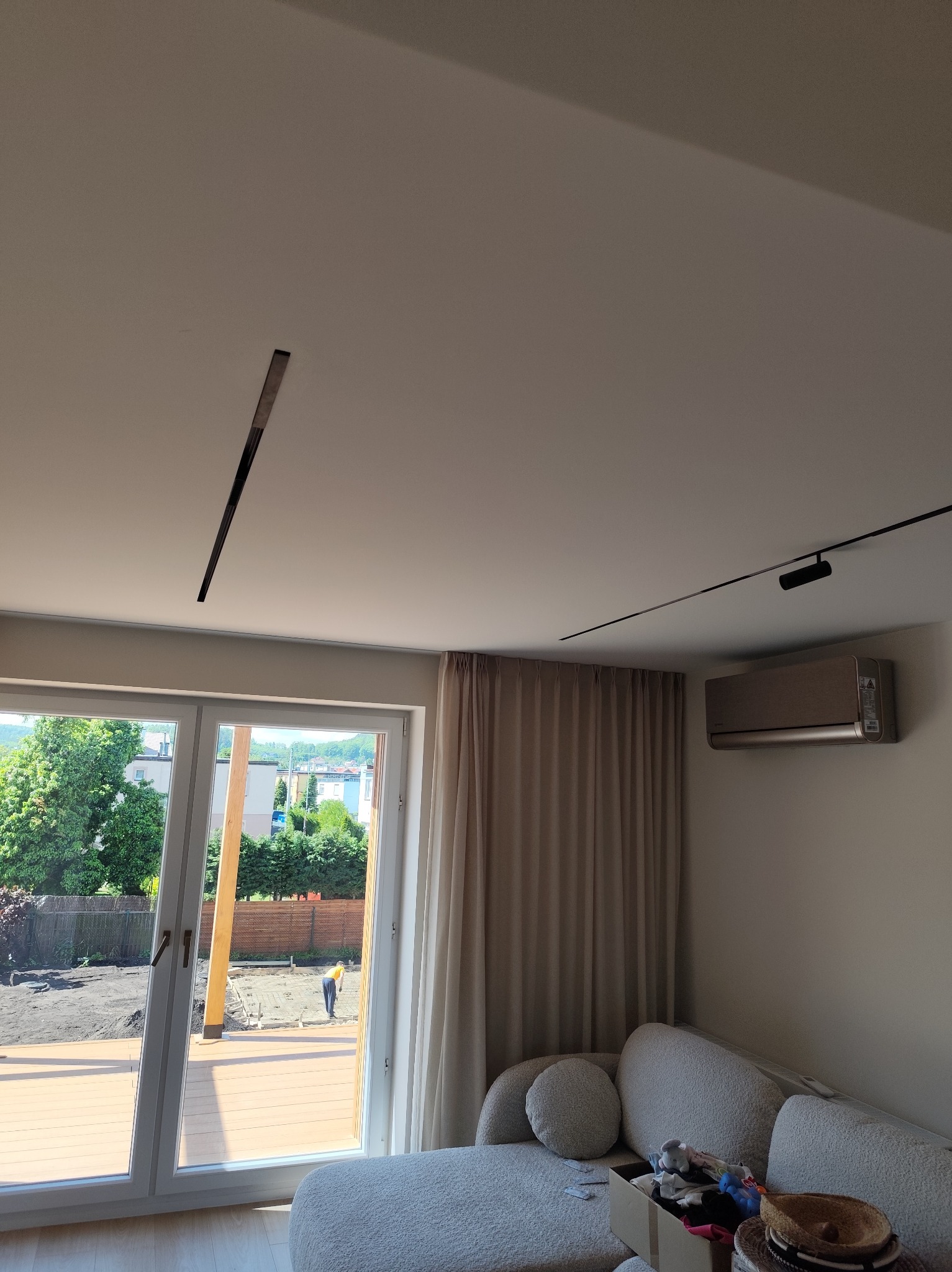 Minimalistyczne wnętrze z oknem na ogród. Na suficie zamontowane nowoczesne oświetlenie liniowe i reflektor. Na ścianie klimatyzator. Wnętrze doświetlone naturalnym światłem.