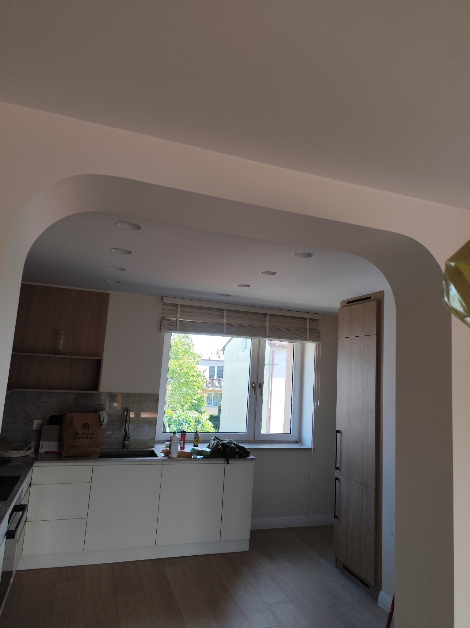 Jasna kuchnia z białymi szafkami i drewnianymi akcentami, widziana przez łukowe przejście. Okno z widokiem na zieleń. Nowoczesny design.