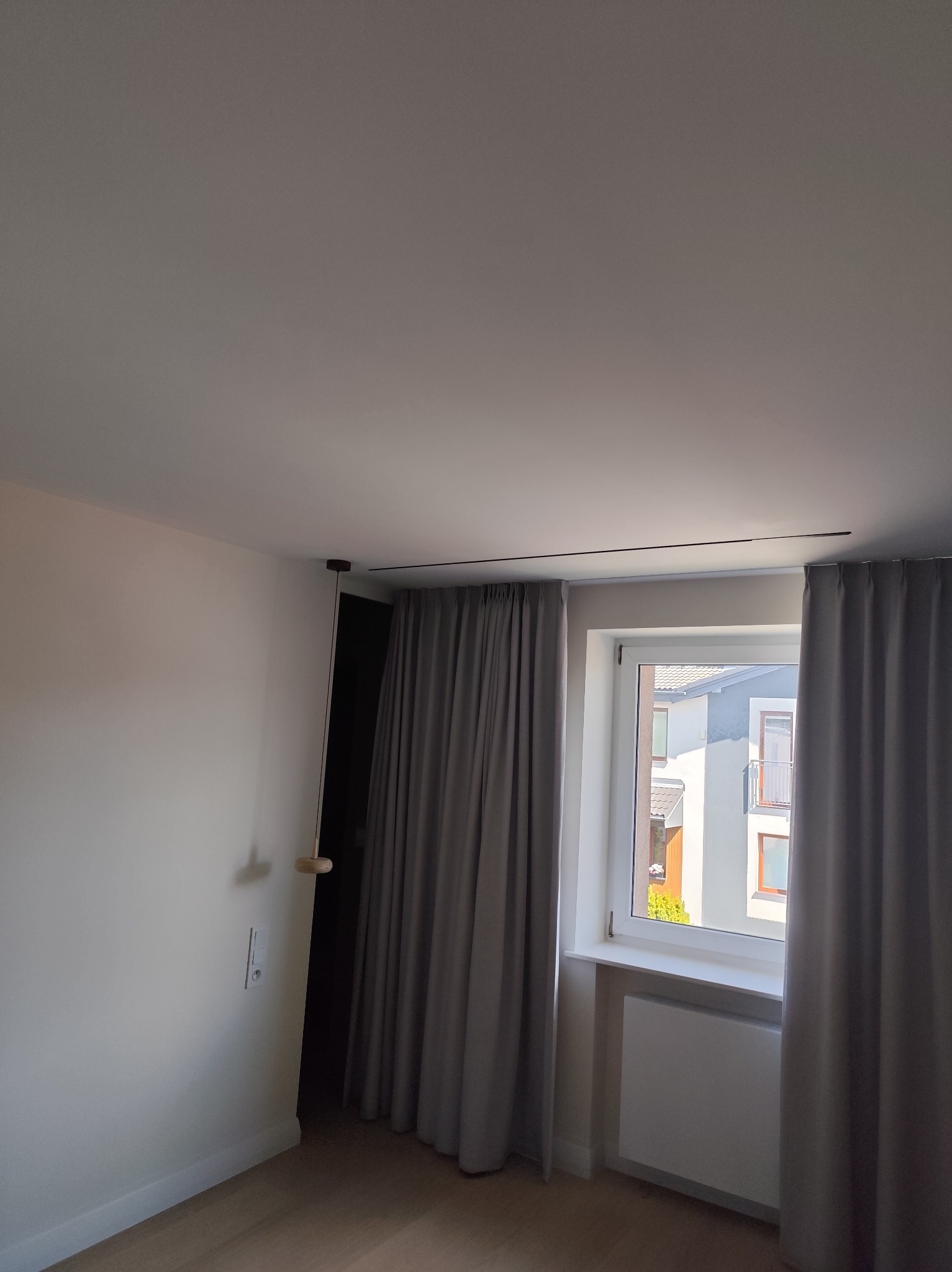 Minimalistyczne wnętrze: szare zasłony na karniszu przy oknie, białe ściany, drewniana podłoga i designerska lampa wisząca. Widok na budynek za oknem.