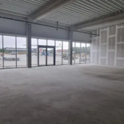 Wnętrze biura w stanie deweloperskim, widoczne ściany przygotowane pod malowanie, duże okna z widokiem na plac budowy. Surowy beton na podłodze.