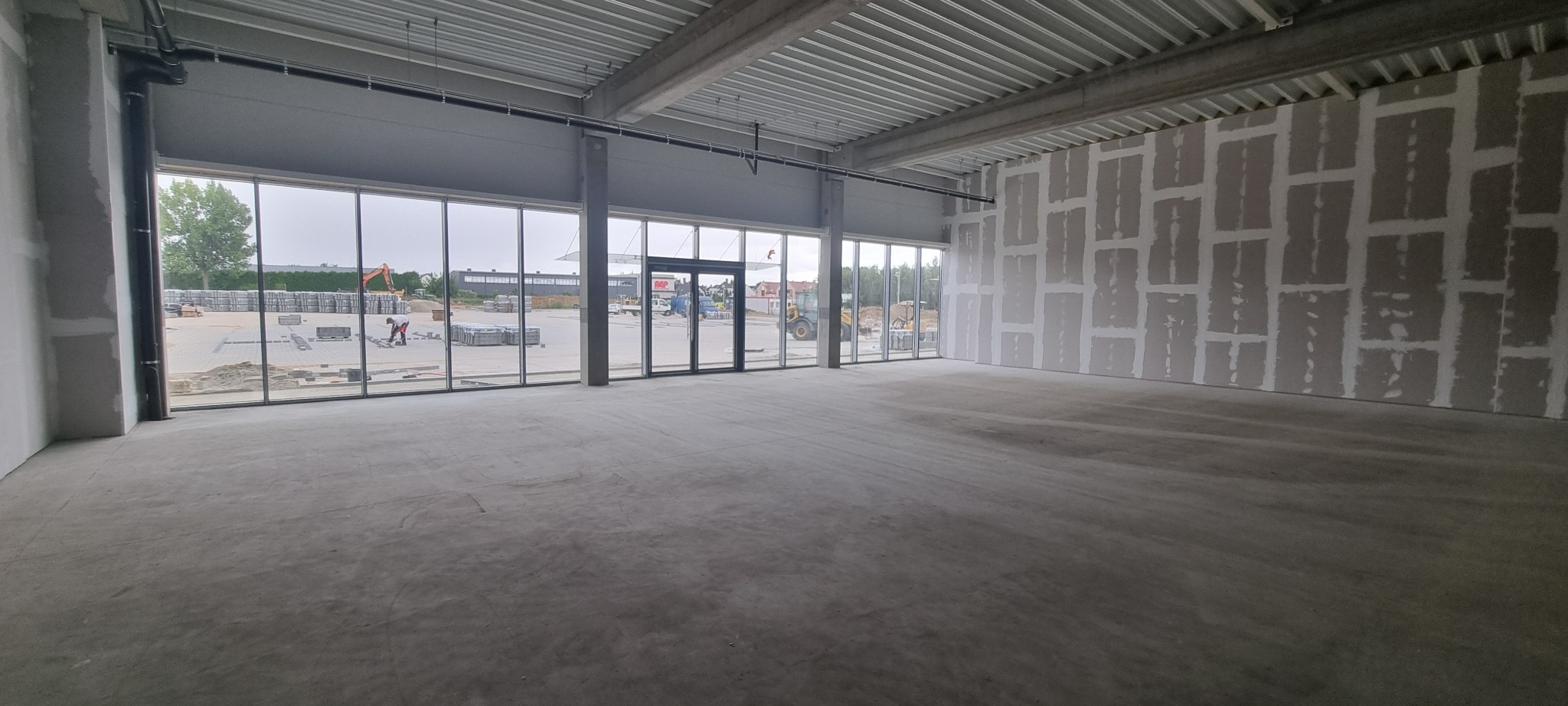 Wnętrze biura w stanie deweloperskim, widoczne ściany przygotowane pod malowanie, duże okna z widokiem na plac budowy. Surowy beton na podłodze.