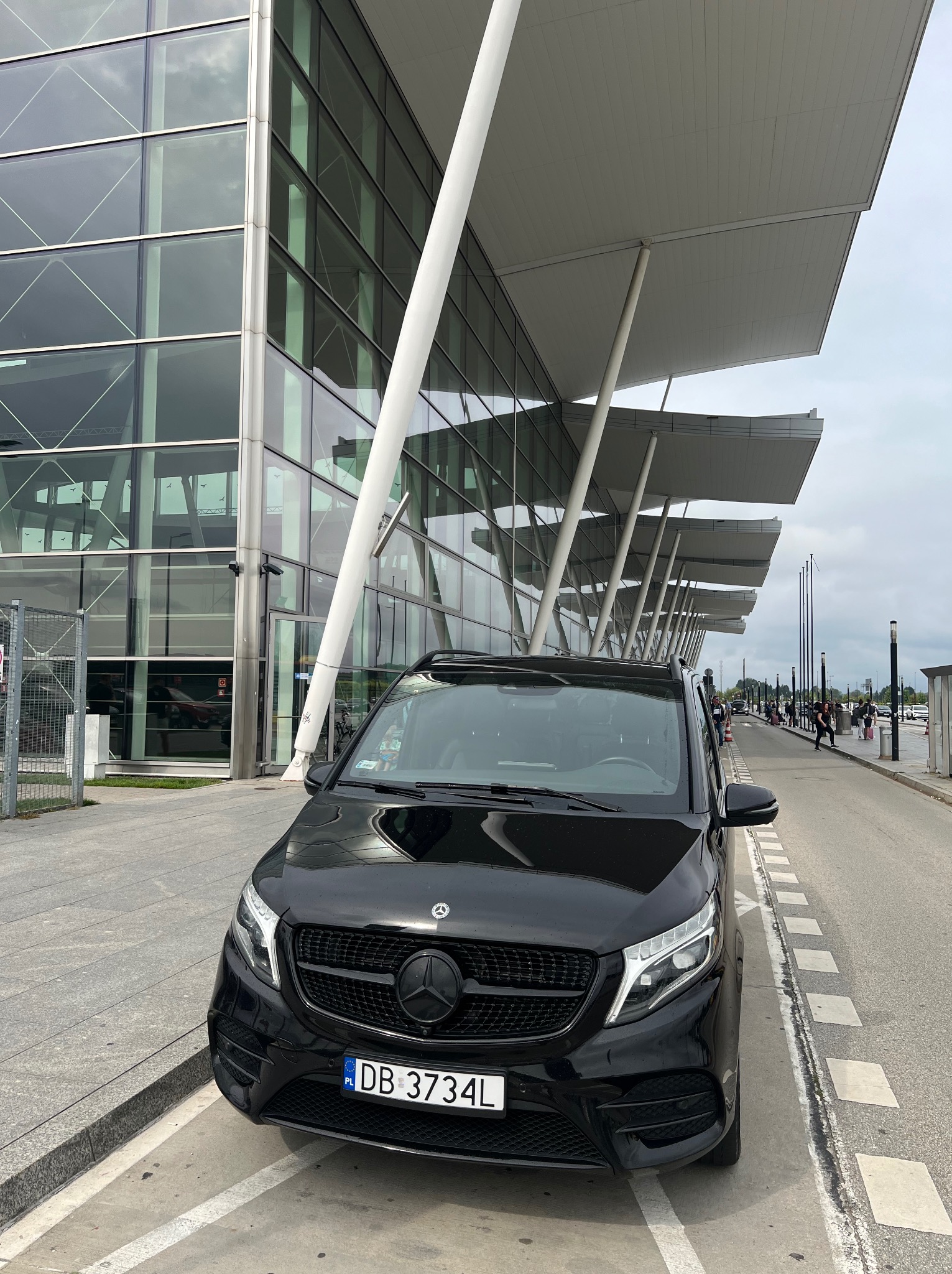 Czarny, luksusowy van marki Mercedes zaparkowany przed nowoczesnym budynkiem terminalu lotniczego z charakterystycznym, wspartym na kolumnach zadaszeniem.