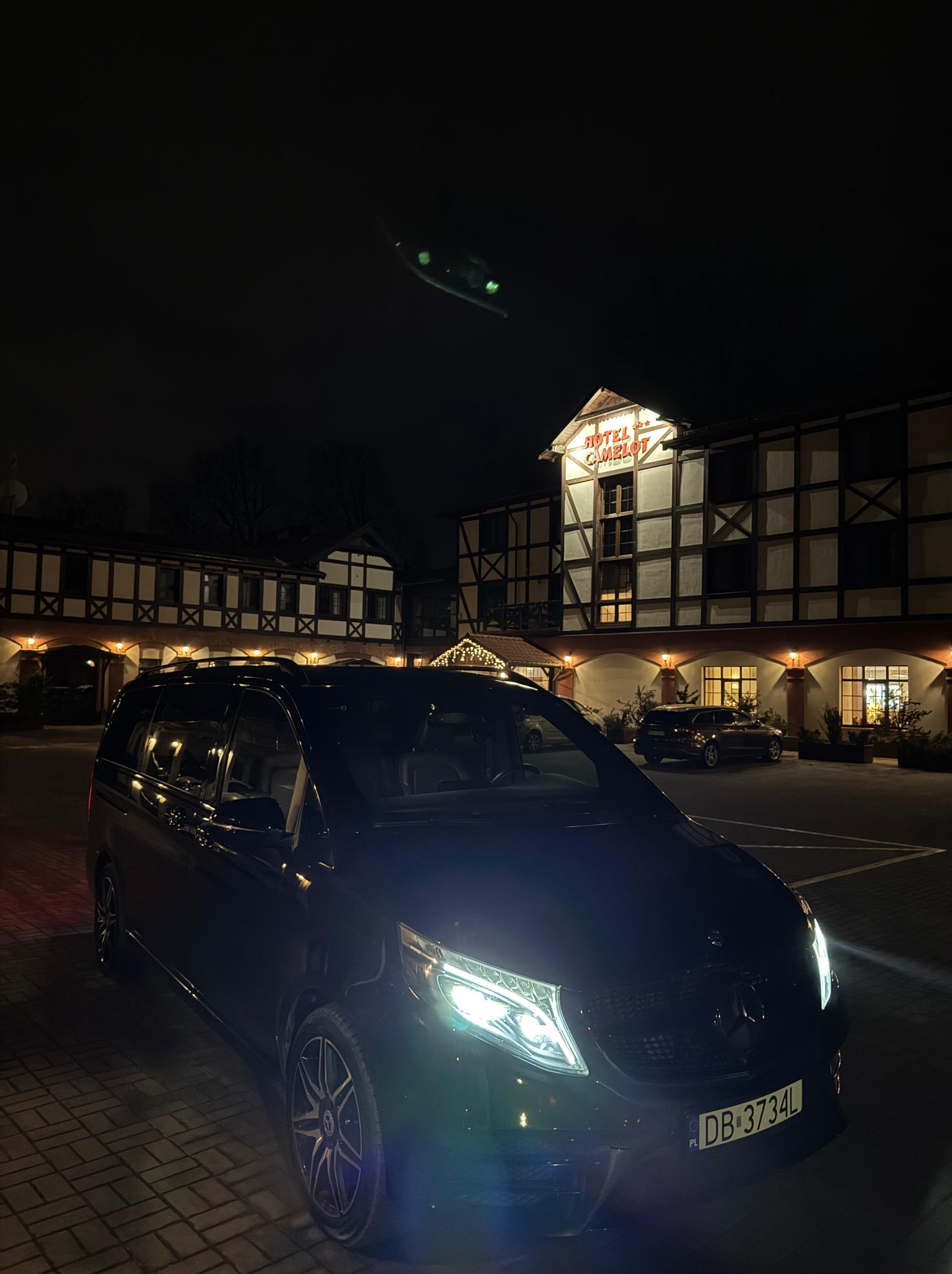 Czarny, luksusowy van Mercedes-Benz V-klasa zaparkowany nocą przed hotelem Camelot z oświetloną fasadą.