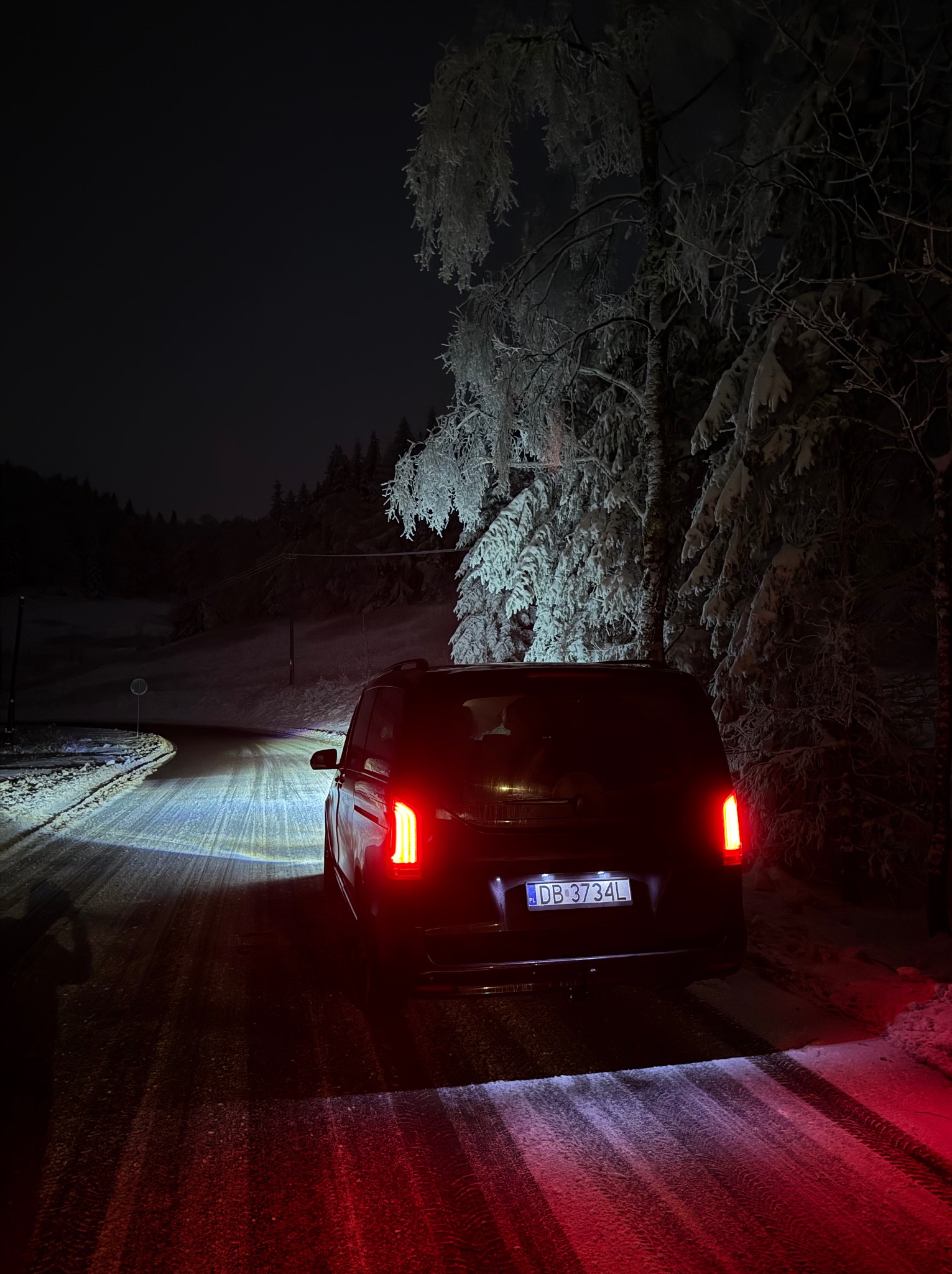 Czarny bus z włączonymi światłami tylnymi, stojący nocą na ośnieżonej drodze, z drzewami pokrytymi szronem w tle i widoczną tablicą rejestracyjną DB 3734L.