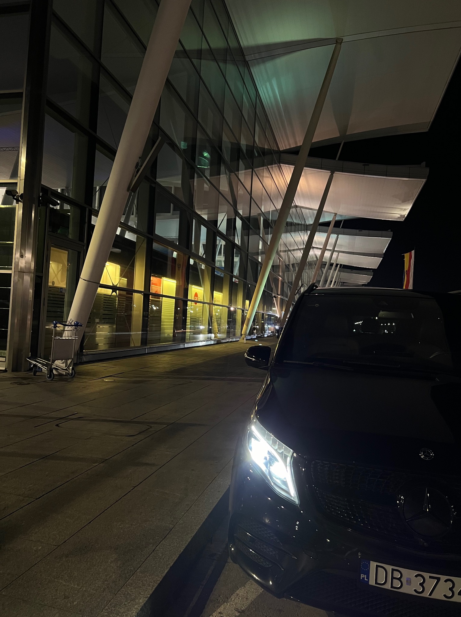 Czarny van Mercedes V-klasy z włączonymi światłami przed lotniskiem w nocy, widoczny fragment terminalu z białymi słupami i szklaną fasadą, w tle wózek bagażowy.