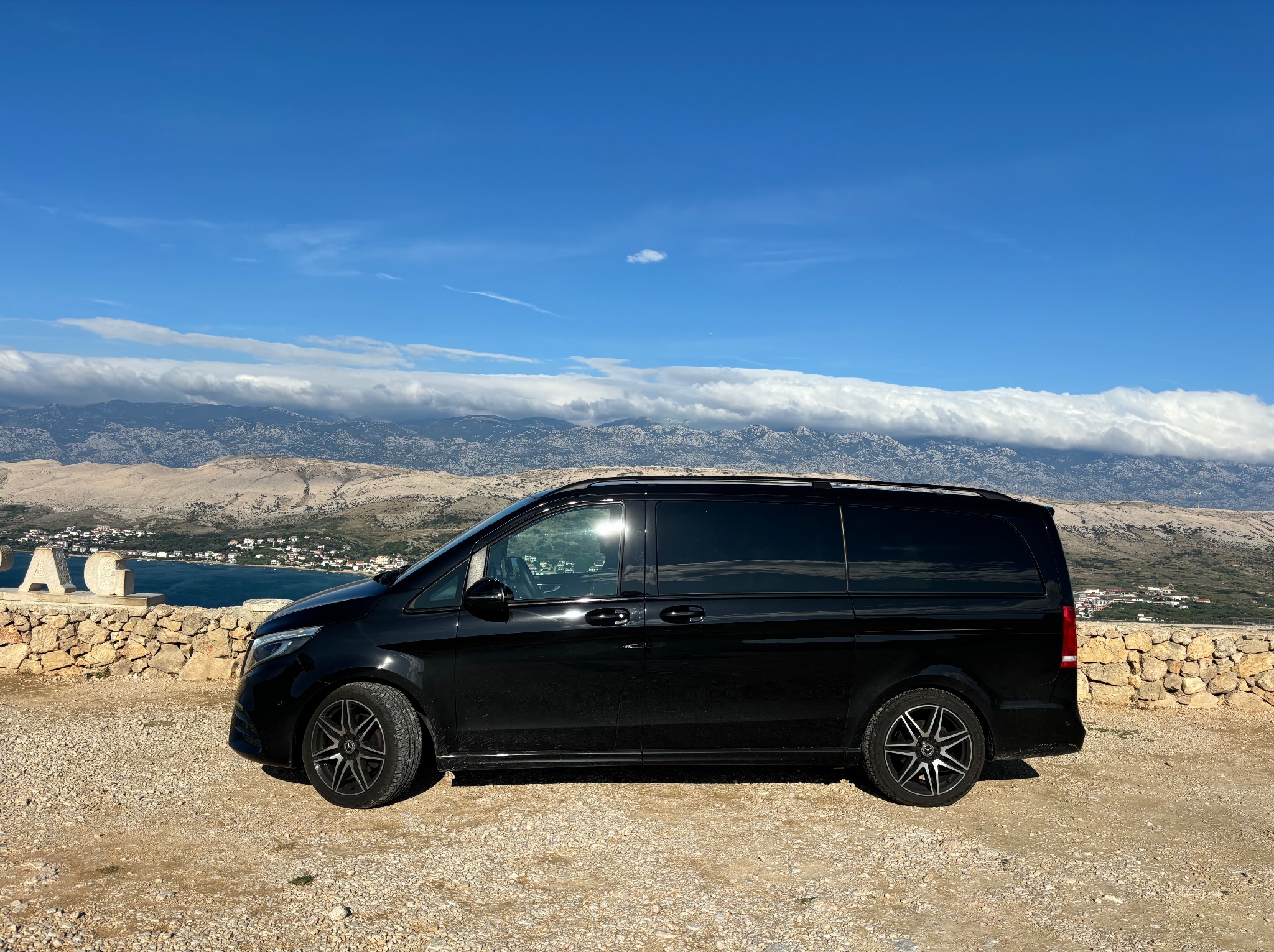 Czarny Mercedes-Benz Vito Tourer zaparkowany na żwirowym wzgórzu z widokiem na morze i góry pod błękitnym niebem z lekkimi obłokami.