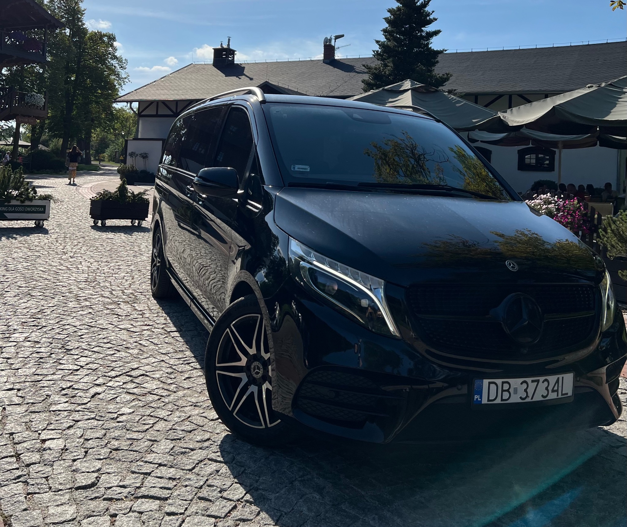 Czarny Mercedes-Benz V-klasy zaparkowany na brukowanej drodze przed budynkiem w stylu dworku z parasolami restauracyjnymi w tle.