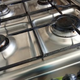 Idea Snails & Garden Daniel Zawrot - Metalowa kuchenka gazowa z czterema palnikami, widoczne czarne ruszty i pokrętła. Powierzchnia płyty jest czysta i błyszcząca, co sugeruje niedawne sprzątanie.