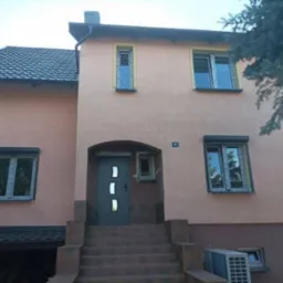Dwupiętrowy dom z beżową elewacją, szarą stolarką okienną i drzwiami wejściowymi, częściowo ocieplony styropianem po prawej stronie, widoczne schody wejściowe i klimatyzator zewnętrzny.