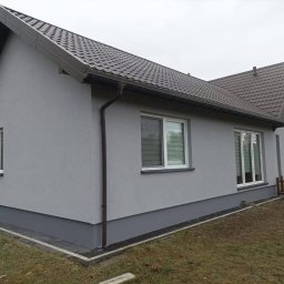 STOCKBUT Amelia Stocka - Dom jednorodzinny po tynkowaniu elewacji na szaro, z widocznymi oknami z roletami i brązowym dachem, cokół w ciemniejszym odcieniu szarości, widoczne obrzeże z kostki.