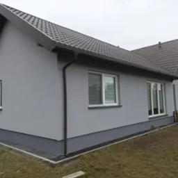 Dom jednorodzinny po tynkowaniu elewacji na szaro, z widocznymi oknami z roletami i brązowym dachem, cokół w ciemniejszym odcieniu szarości, widoczne obrzeże z kostki.