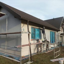 STOCKBUT Amelia Stocka - Dwóch pracowników na rusztowaniu maluje elewację domu jednorodzinnego w jasnych barwach, okna zabezpieczone folią, widoczny fragment dachu z brązową dachówką.