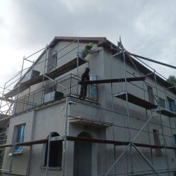 STOCKBUT Amelia Stocka - Dwóch pracowników na rusztowaniu podczas prac elewacyjnych przy dwupiętrowym domu z balkonem, jeden maluje ścianę pod dachem, drugi stoi na balkonie z wiadrem farby.