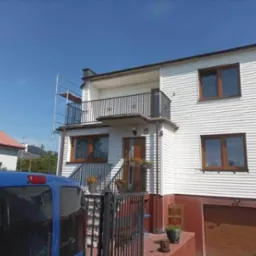 Dom z elewacją z białego sidingu, balkonem z czarną balustradą i częściowo pomalowanym na czerwono cokołem, widoczne rusztowanie z boku budynku.