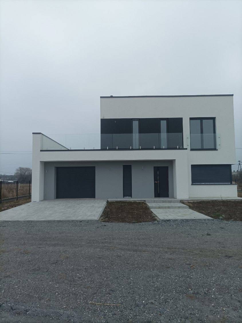 Nowoczesny dwukondygnacyjny dom z płaskim dachem, szarą elewacją i balkonem z szklaną balustradą, widoczne okna z roletami zewnętrznymi, garaż z bramą segmentową i chodnik z kostki brukowej.