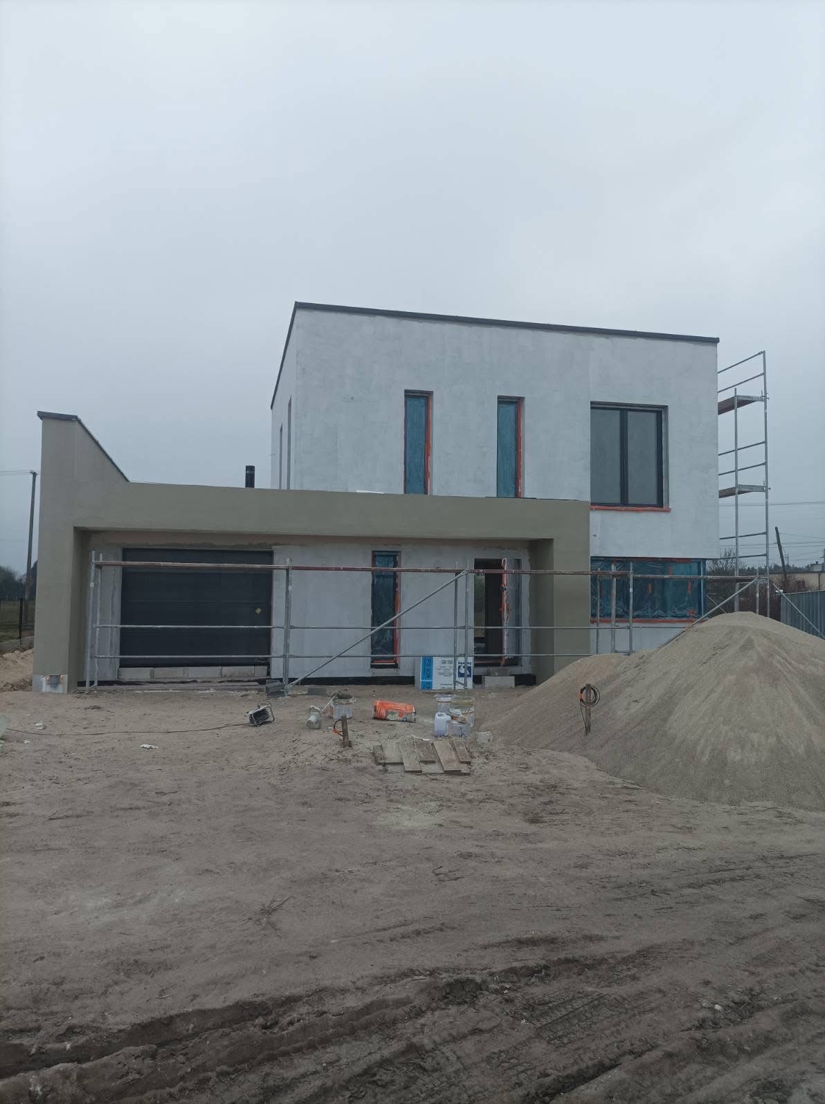 Nowoczesny dom jednorodzinny w trakcie prac elewacyjnych: świeżo otynkowana fasada, rusztowania, zabezpieczone okna folią, piasek budowlany na posesji.