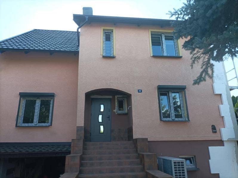 Dwupiętrowy dom z beżową elewacją, szarą stolarką okienną i drzwiami wejściowymi, częściowo ocieplony styropianem po prawej stronie, widoczne schody wejściowe i klimatyzator zewnętrzny.