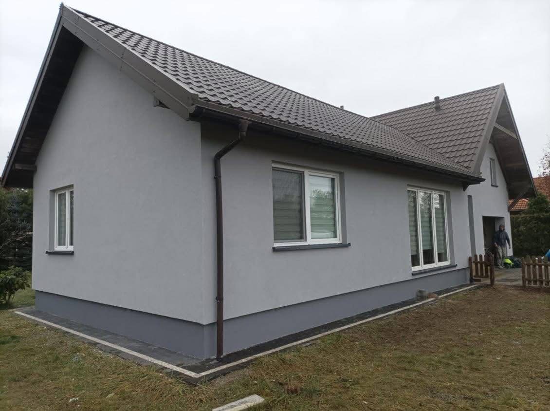 Dom jednorodzinny po tynkowaniu elewacji na szaro, z widocznymi oknami z roletami i brązowym dachem, cokół w ciemniejszym odcieniu szarości, widoczne obrzeże z kostki.