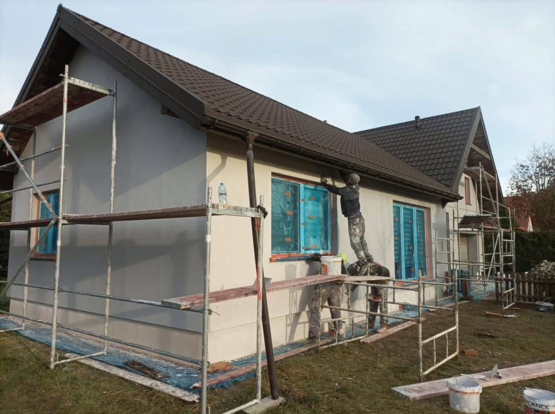 Dwóch pracowników na rusztowaniu maluje elewację domu jednorodzinnego w jasnych barwach, okna zabezpieczone folią, widoczny fragment dachu z brązową dachówką.