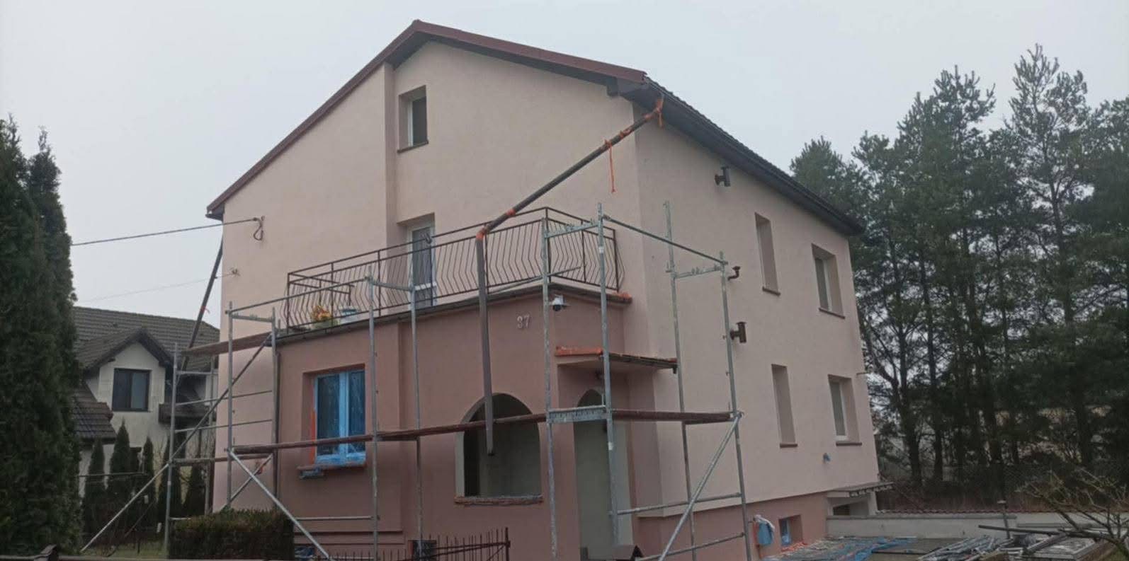 Dwukondygnacyjny dom w trakcie prac elewacyjnych, z rusztowaniami i zabezpieczonym oknem, widoczny przewód do podawania materiału.