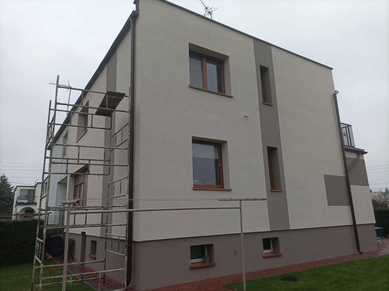 Dwukondygnacyjny dom z jasnoszarą elewacją i brązowymi akcentami, widoczne rusztowanie i fragment suszarki na pranie.