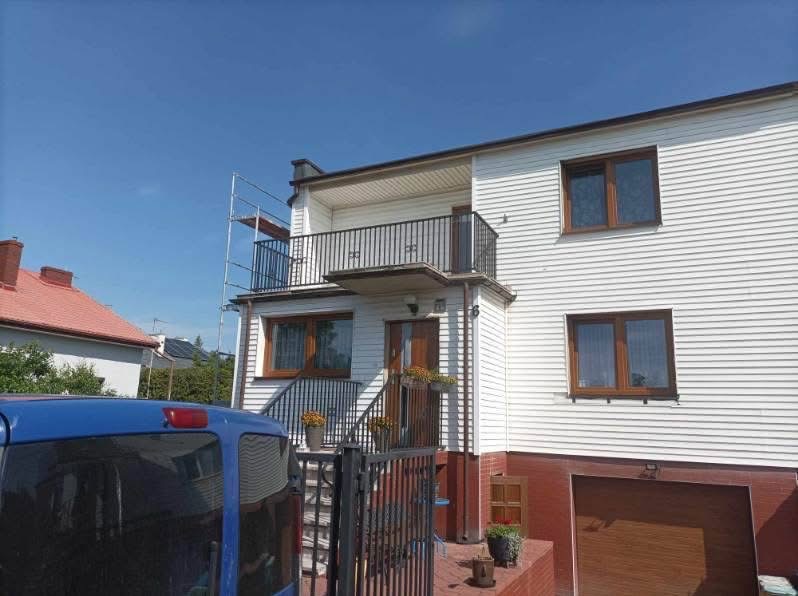 Dom z elewacją z białego sidingu, balkonem z czarną balustradą i częściowo pomalowanym na czerwono cokołem, widoczne rusztowanie z boku budynku.