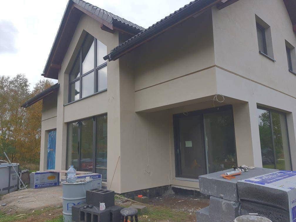 Dwukondygnacyjny dom jednorodzinny z jasną elewacją w trakcie prac wykończeniowych, widoczne materiały budowlane i narzędzia na placu budowy.