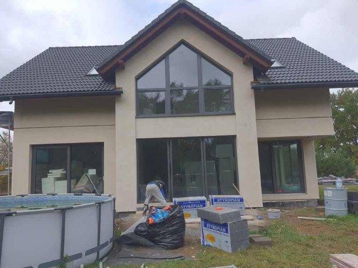 Dwukondygnacyjny dom jednorodzinny z ocieploną elewacją w trakcie prac wykończeniowych, widoczne paczki styropianu, pracownik w roboczym ubraniu i basen ogrodowy.