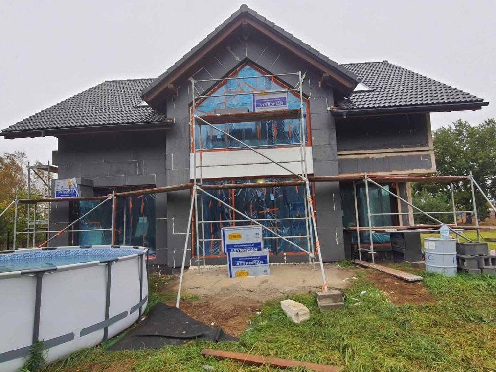 Dom jednorodzinny w trakcie ocieplania, widoczne rusztowania, styropianowe płyty na elewacji, okna zabezpieczone folią, obok basen ogrodowy.