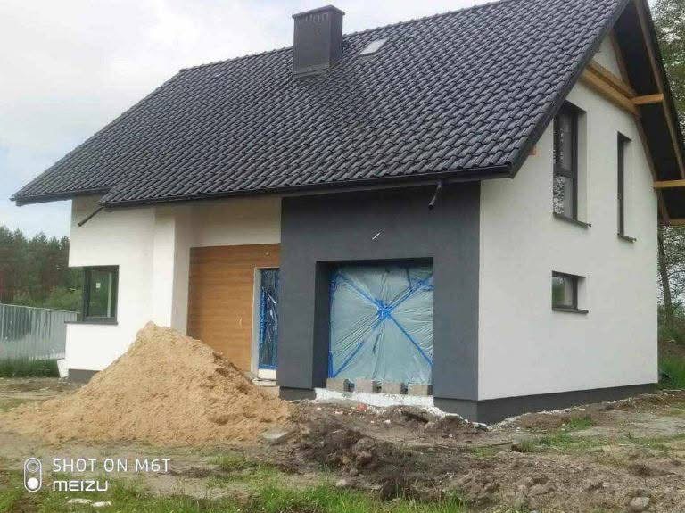 Nowoczesny dom jednorodzinny w trakcie wykańczania elewacji: fragment otynkowanej ściany pomalowanej na biało i szaro, okno zabezpieczone folią, sterta piasku przed budynkiem.