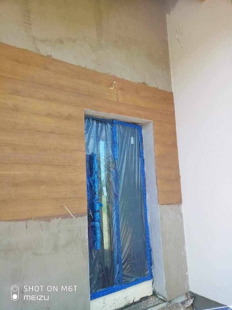 Montaż drewnopodobnej elewacji poziomej na fragmencie ściany zewnętrznej, okno zabezpieczone folią ochronną, widoczne niedokończone prace wykończeniowe.